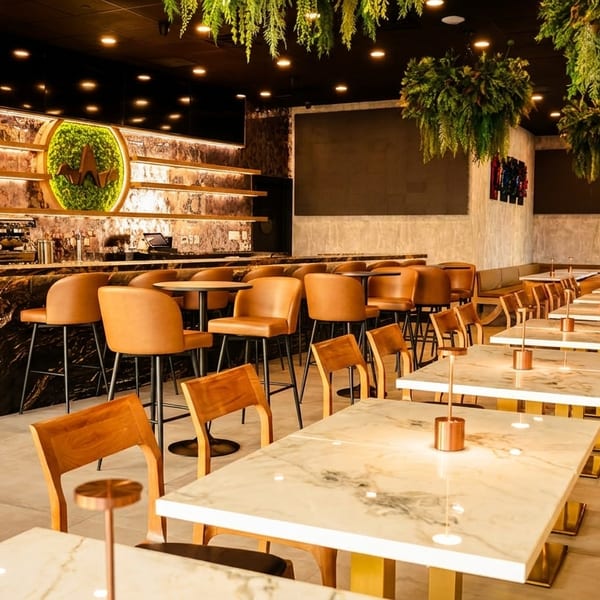 Modern restaurant interior with a marble bar, brown leather stools, and white marble tables with gold accents.