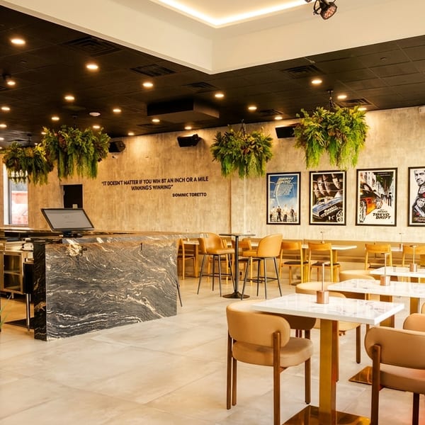 Modern restaurant interior with marble bar, light wood tables, tan chairs, and movie posters on the wall.