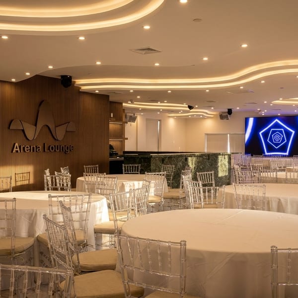 Arena Lounge event space with round tables, clear chairs, and a large screen displaying a blue geometric pattern.