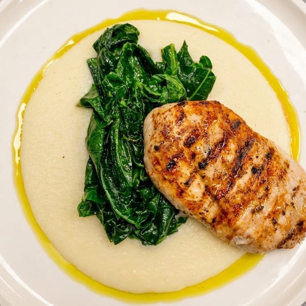 Grilled chicken breast with sautéed greens and polenta, drizzled with olive oil on a white plate.