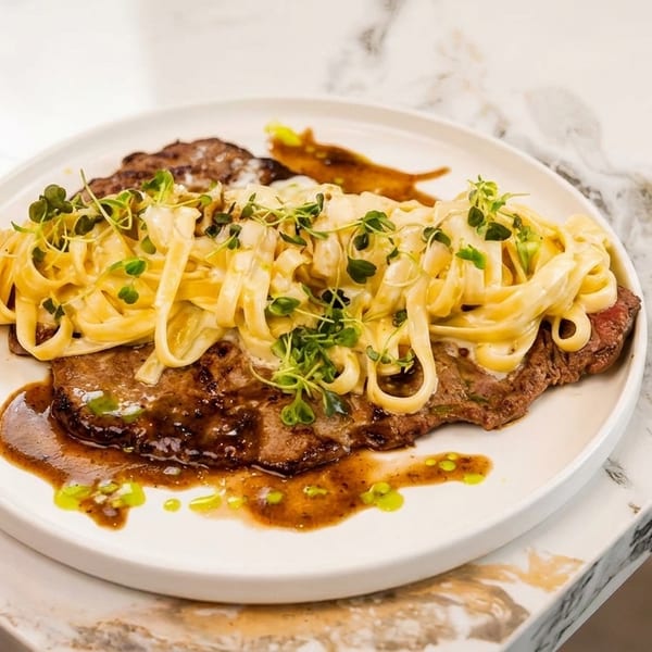 Steak with fettuccine alfredo and microgreens on a white plate, on a marble counter.